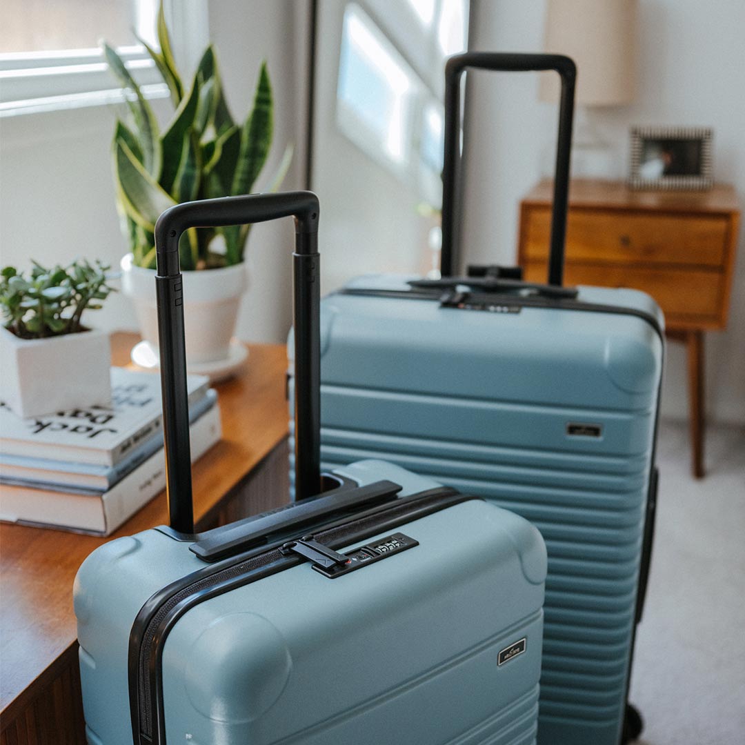 The Arlo Skye Zipper Carry-On Max and Zipper Check-In in Sea Sage. Both bags are standing in the bedroom.