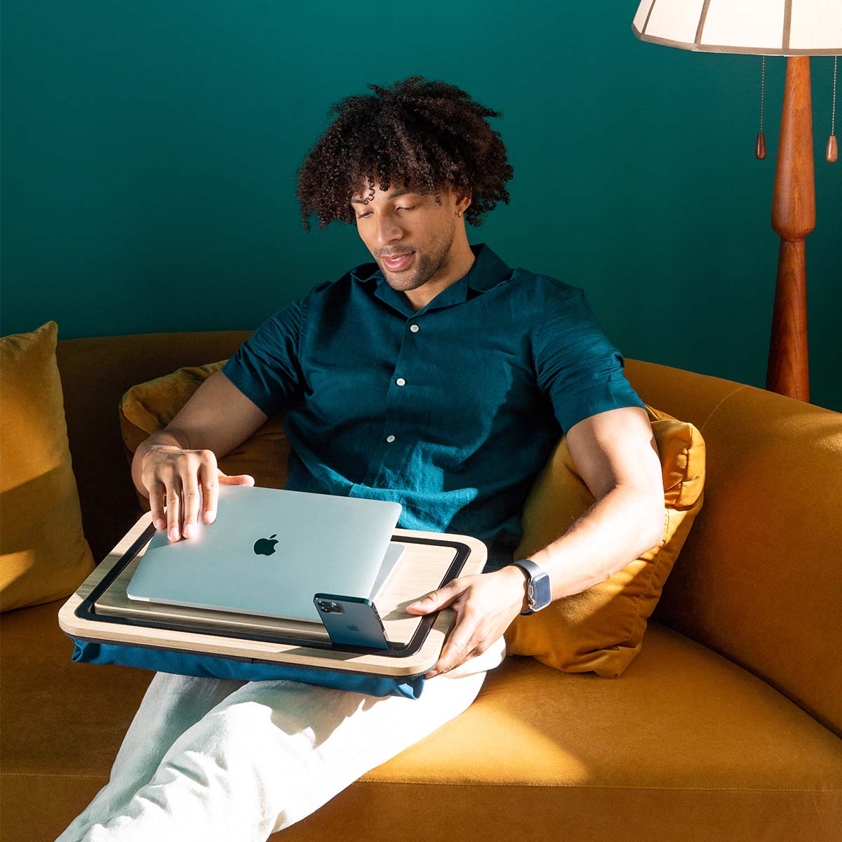 man sitting on sofa with lap desk on lap and man opening laptop while phone is mounted on the stand.