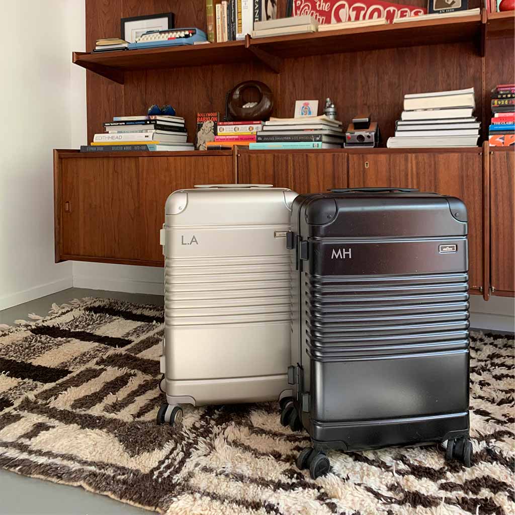 Two Arlo Skye Frame Carry-On Max suitcases in Black and Champagne color with initials laser engraved on the top left side of the carry-ons. Initials on champagne carryon say "LA" and initials on Black Carry-On say "MH"