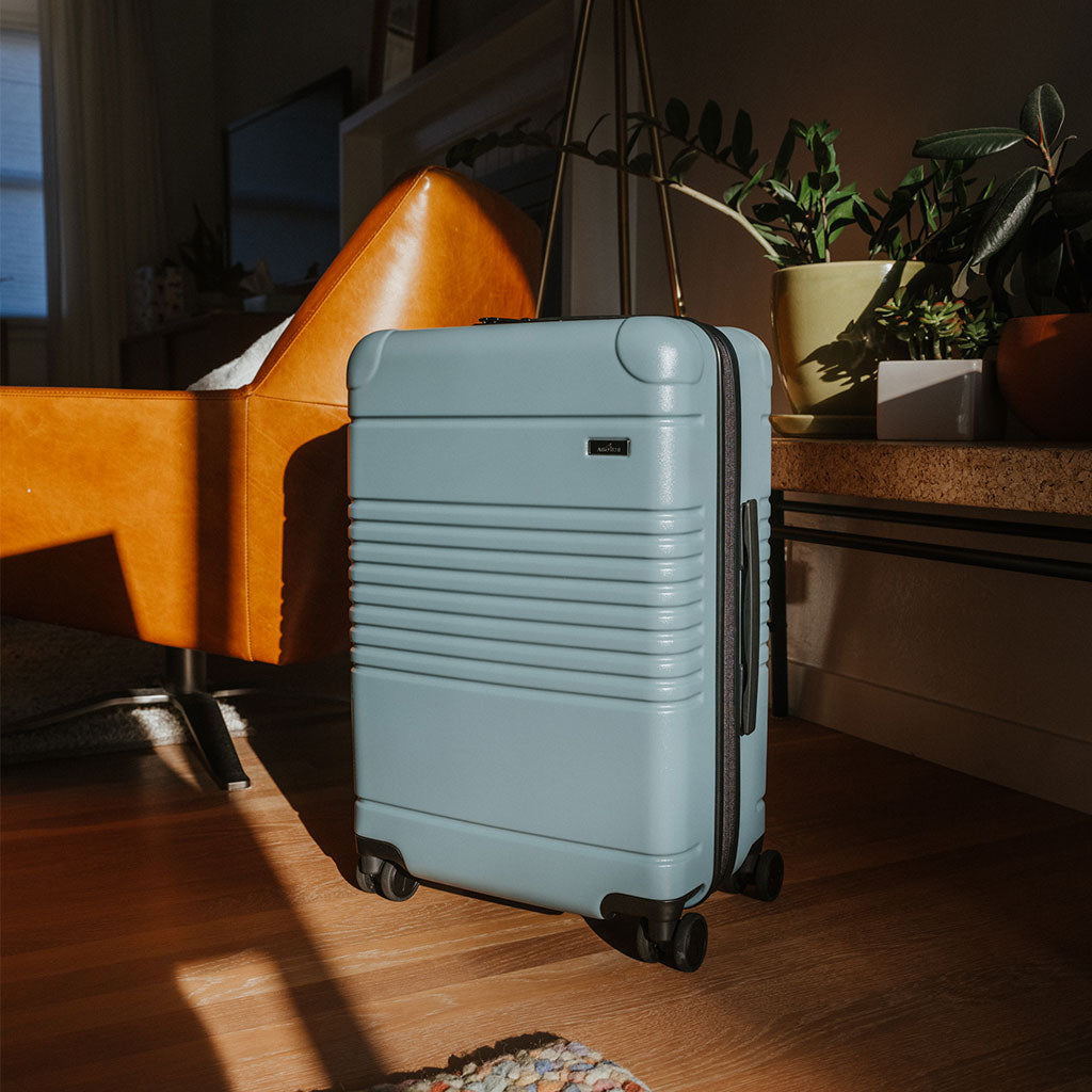 An Arlo Skye Zipper Carry-On Max in Sea Sage inside a living room of a home with some sunlight streaming in and hitting the carry-on.