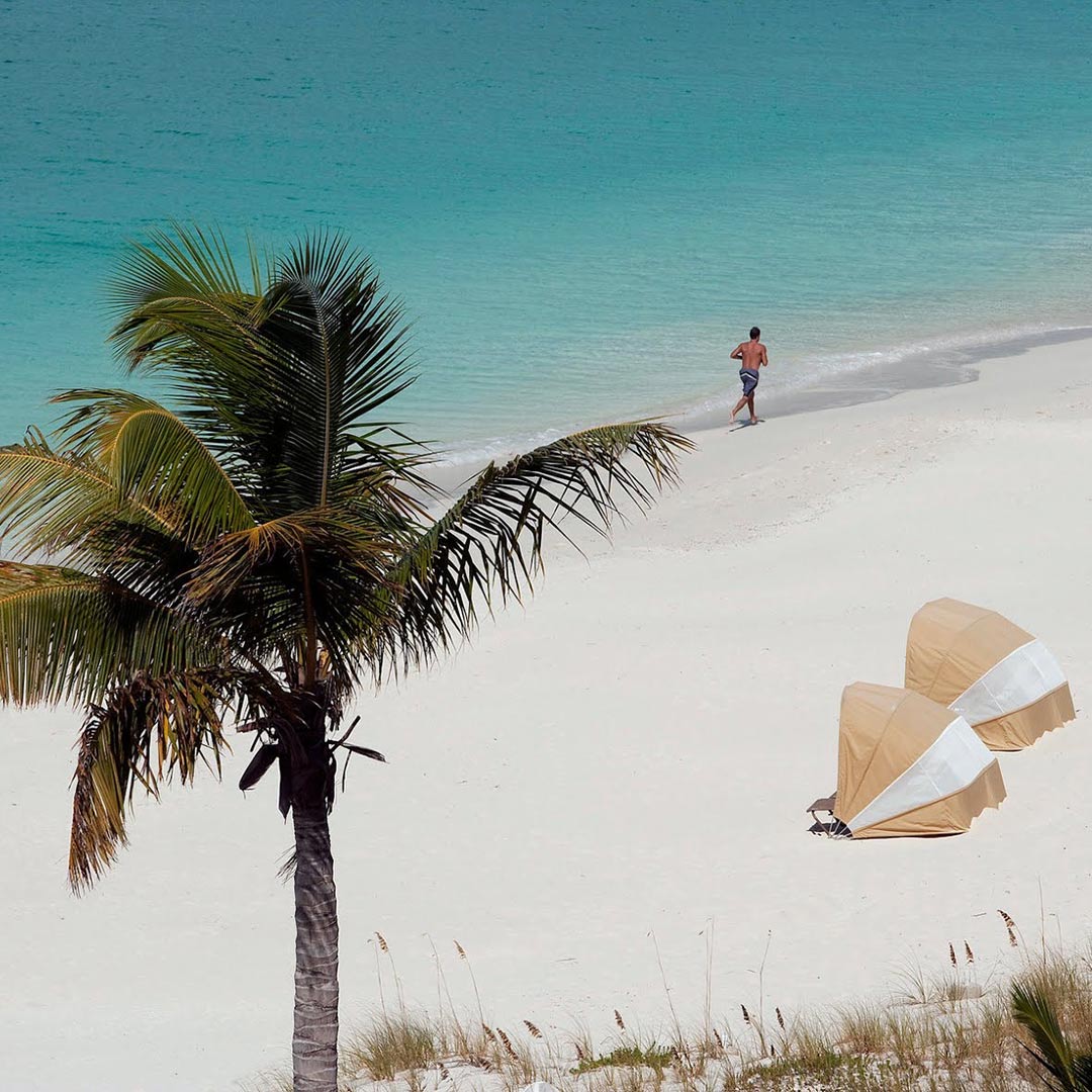 A few of the beach in the Caribbean with 2 yellow sunbeds, a man by the water and a palm tree.