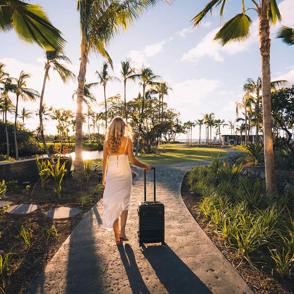 A girl in white dress with black Arlo Skye Frame Carry-On Max walking down a pathway in a lush tropical garden.