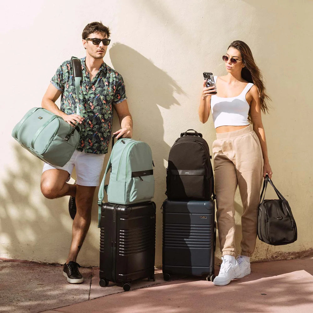 boy and girl standing with 2 carry-ons in black and blue and 2 back packs on carry-ons in mint and black and holding 2 weekenders in mint and black. 